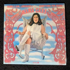 Könnte beinhalten: Albumcover für "Bubble Gum, Lemonade &... Something for Mama" von Mama Cass. Das Cover zeigt eine Frau in weißem Kleid und Stiefeln, die in einem Korbstuhl vor einem blauen Himmel mit rosa Dekorationselementen sitzt.