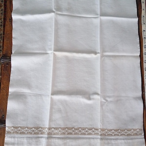 May include: A rectangular white kitchen towel featuring a textured weave and a decorative tan lace border along the bottom edge. A white measuring tape is visible on the right side, indicating the towel measures approximately 13 inches in width.