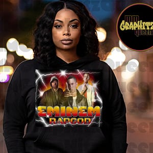 May include: Black hoodie with a graphic print of Eminem, with the text "Eminem Rapgod" in white letters. The graphic features Eminem and two other men in a stylized, comic-book style.