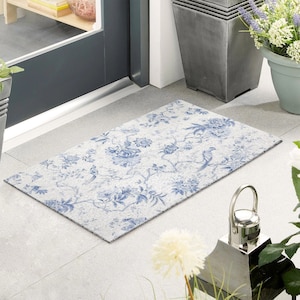 Puede incluir: Una alfombra blanca para la entrada con un patrón floral azul y un diseño de pájaro. La alfombra está hecha de un material texturizado y se coloca delante de una puerta.