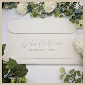 May include: A cream-coloured pregnancy notes holder, featuring the text "Baby William's PREGNANCY NOTES" in gold script. The rectangular holder has a flap closure. White roses and greenery are arranged around it.