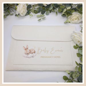 May include: Cream-coloured pregnancy notes holder with a flap closure. A pocket on the front features a sleeping bunny illustration and the text "Baby Evans Pregnancy Notes" in gold script. The holder is surrounded by white roses and greenery.