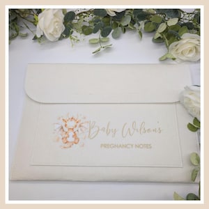 May include: Cream-coloured pregnancy notes holder with a flap closure. The front pocket features a watercolour illustration of a baby dragon and the text "Baby Wilson's Pregnancy Notes" in gold script. White roses and greenery surround the holder.