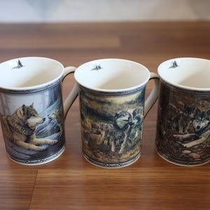 May include: Three ceramic mugs featuring detailed illustrations of wolves in natural habitats. Each mug has a cream-colored interior with a small wolf icon near the rim, a curved handle, and a decorative border pattern at the top and bottom edges.