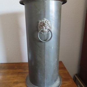 May include: A tall, silver metal umbrella stand with a lion head design and a ring handle. The stand has a round base and a cylindrical body.
