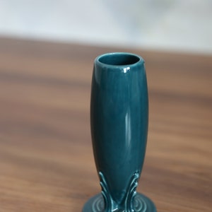 May include: A teal ceramic bud vase with a smooth, glossy finish. The vase features a cylindrical body that tapers slightly toward the top and flares out at the base. The base is decorated with raised, symmetrical, fan-like molded details.
