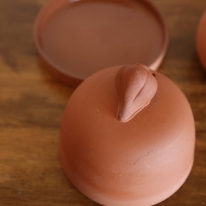 May include: A terracotta lid with a small knob on top. The lid is slightly rounded and has a smooth surface.