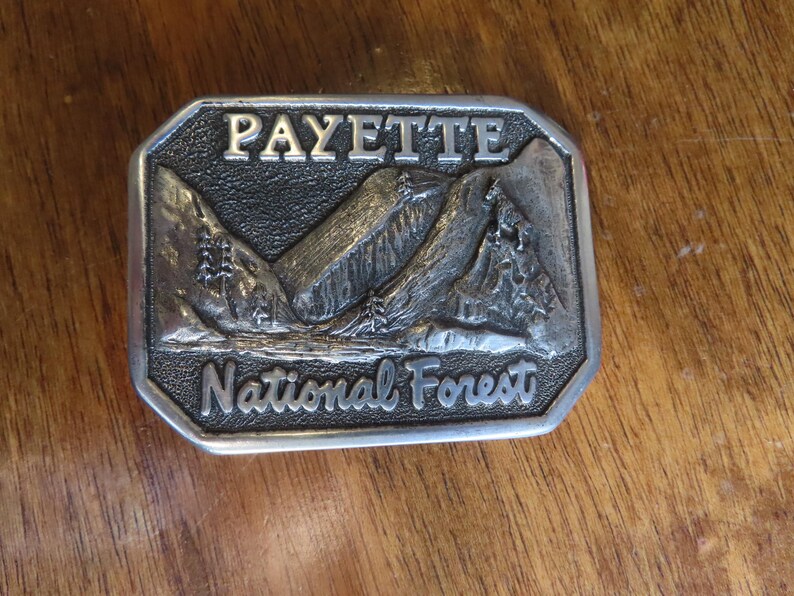 Op de afbeelding: Zilveren gesp met een reli&euml;fbeeld van bergen en bomen. De gesp heeft de tekst "Payette National Forest".
