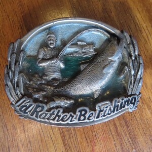 Op de afbeelding: Een zilveren gesp met een groene en blauwe achtergrond. Een man vist met een hengel en reel. Een grote vis springt uit het water. De gesp zegt "I'd Rather Be Fishing".