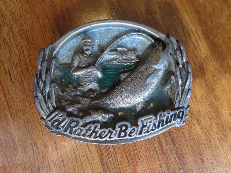 Op de afbeelding: Een zilveren gesp met een groene en blauwe achtergrond. Een man vist met een hengel en reel. Een grote vis springt uit het water. De gesp zegt "I'd Rather Be Fishing".