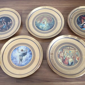 May include: A set of five round decorative plates featuring gold-toned rims with intricate patterns and central circular illustrations depicting various religious scenes, including figures in robes, a nativity scene, and a figure ascending into clouds.