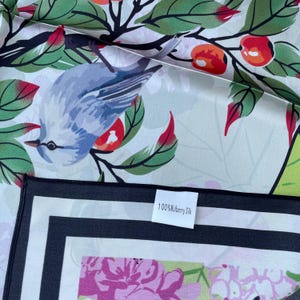 May include: A silk scarf featuring a light blue bird perched on branches with green leaves and orange berries. The fabric has a white background and a black border. A white tag attached to the edge displays the text 100% Mulberry Silk.
