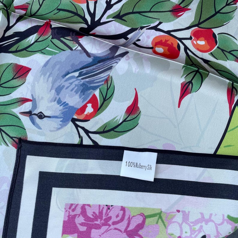 May include: A silk scarf featuring a light blue bird perched on branches with green leaves and orange berries. The fabric has a white background and a black border. A white tag attached to the edge displays the text 100% Mulberry Silk.