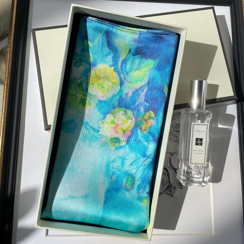 K&ouml;nnte beinhalten: Ein rechteckiges Seidentuch mit einem lebendigen Blumenmuster im Aquarellstil in Blau-, T&uuml;rkis- und Gelbt&ouml;nen, pr&auml;sentiert in einer wei&szlig;en Geschenkbox neben einer klaren Glasflasche Jo Malone Red Roses K&ouml;lnisch Wasser mit silbernem Verschluss.