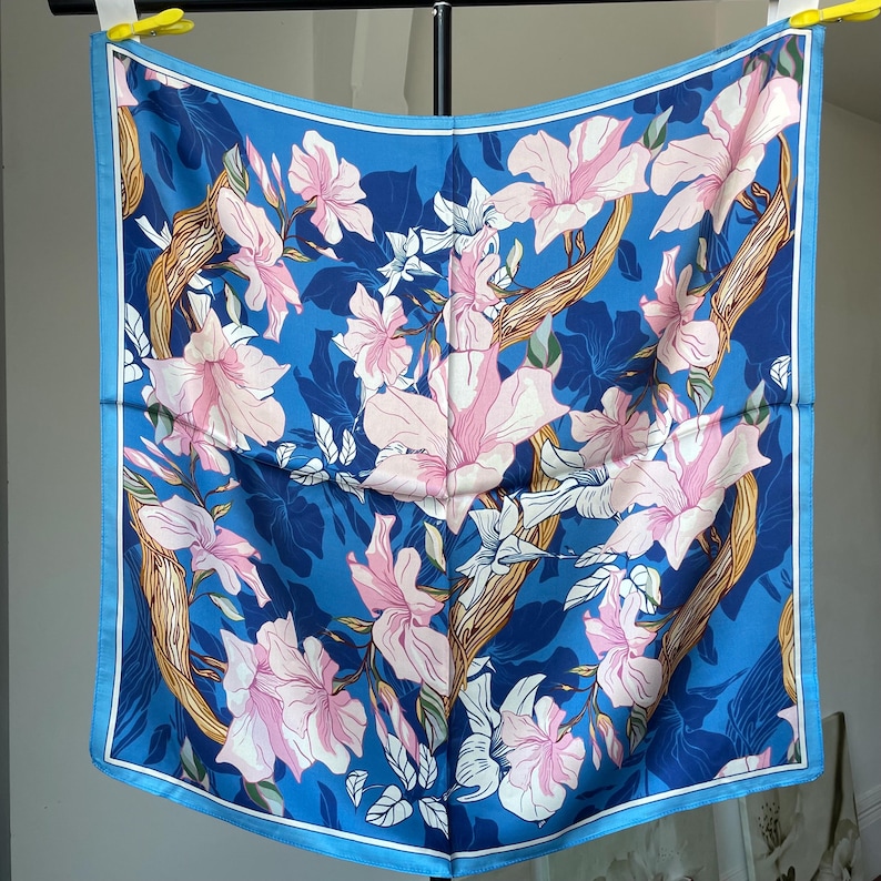 May include: A square scarf featuring a vibrant blue background with a repeating pattern of light pink flowers and tan, textured tree branches. The design includes dark blue leaf silhouettes and a thin light blue border framing the entire square piece.