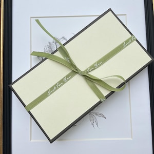 May include: A rectangular gift box with a cream-colored lid and dark brown sides, wrapped in a sage green ribbon tied in a bow. The ribbon features the repeating white text "Just For You" printed along its length in a cursive script font.