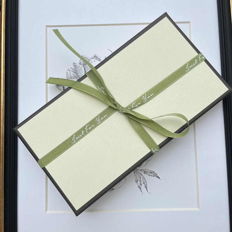 May include: A rectangular gift box with a cream-colored lid and dark brown sides, wrapped in a sage green ribbon tied in a bow. The ribbon features the repeating white text "Just For You" printed along its length in a cursive script font.