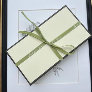 K&ouml;nnte beinhalten: Eine rechteckige Geschenkbox mit cremefarbenem Deckel und dunkelbraunen Seiten, umwickelt mit einem salbeigr&uuml;nen Band, das zu einer Schleife gebunden ist. Das Band tr&auml;gt den wiederkehrenden wei&szlig;en Schriftzug "Just For You" in Schreibschrift.