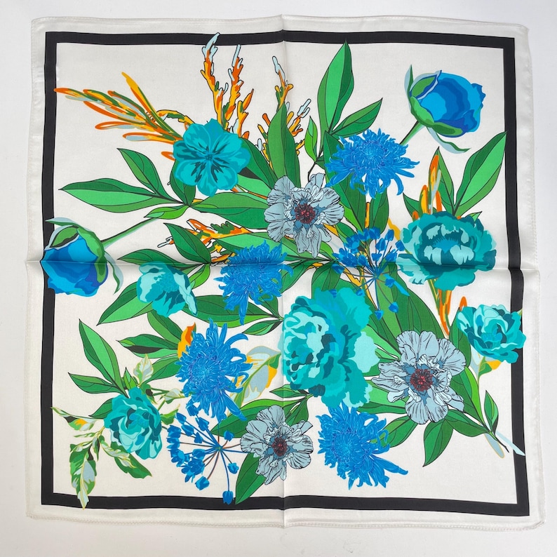K&ouml;nnte beinhalten: Ein quadratisches Tuch mit wei&szlig;em Hintergrund und schwarzem Rand. Die Mitte zeigt ein lebhaftes Blumenarrangement mit verschiedenen Blau- und T&uuml;rkist&ouml;nen, gr&uuml;nen Bl&auml;ttern und d&uuml;nnen orangefarbenen Akzenten im gesamten botanischen Design.