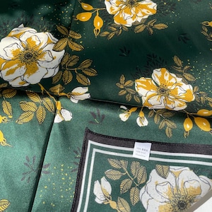 K&ouml;nnte beinhalten: Ein dunkelgr&uuml;nes Seidentuch mit einem sich wiederholenden Muster aus wei&szlig;en und gelben Blumen mit dunkelgr&uuml;nen Bl&auml;ttern. Ein wei&szlig;es Stoffetikett ist an der Ecke befestigt und zeigt den Text 100% 真蚕丝, 100% SILK und 55cmx55cm.