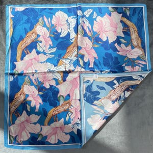 May include: A square scarf featuring a vibrant blue background with a repeating pattern of light pink flowers and brown, textured tree branches. The design includes a thin white border framing the entire piece, with a folded corner showing the reverse.