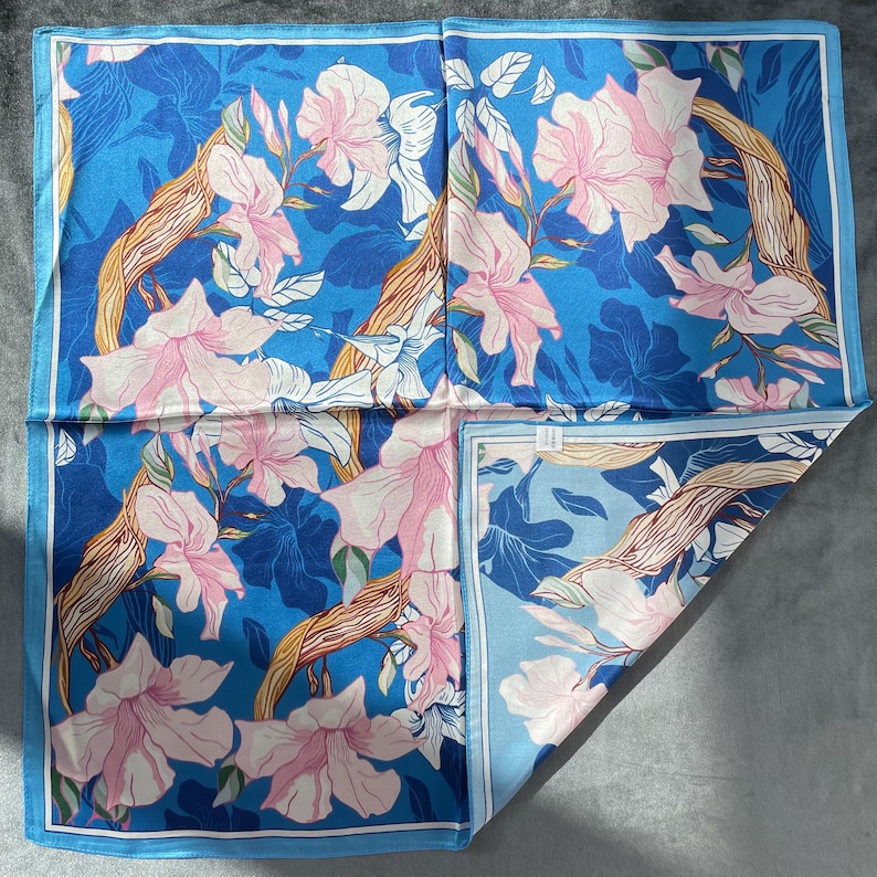 May include: A square scarf featuring a vibrant blue background with a repeating pattern of light pink flowers and brown, textured tree branches. The design includes a thin white border framing the entire piece, with a folded corner showing the reverse.