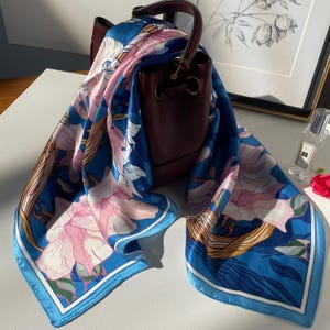 May include: A square silk scarf featuring a vibrant blue background with large, light pink floral patterns and golden brown leaf accents, draped over a dark burgundy leather bucket bag with a structured handle and gold-toned hardware.