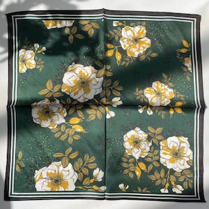 K&ouml;nnte beinhalten: Ein quadratisches Tuch mit dunkelgr&uuml;nem Hintergrund und einem sich wiederholenden Muster aus wei&szlig;en und gelben Blumen, Bl&auml;ttern und Knospen. Das Tuch ist von einer d&uuml;nnen wei&szlig;en Linie und einem schwarzen Rand an allen vier Kanten umgeben.