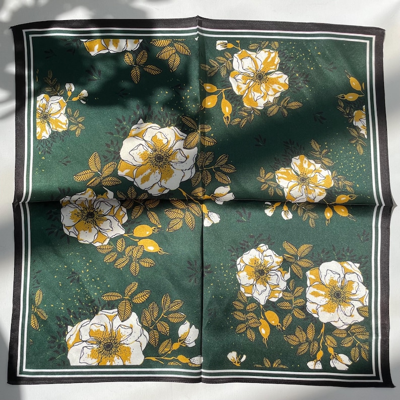 K&ouml;nnte beinhalten: Ein quadratisches Tuch mit dunkelgr&uuml;nem Hintergrund und einem sich wiederholenden Muster aus wei&szlig;en und gelben Blumen, Bl&auml;ttern und Knospen. Das Tuch ist von einer d&uuml;nnen wei&szlig;en Linie und einem schwarzen Rand an allen vier Kanten umgeben.