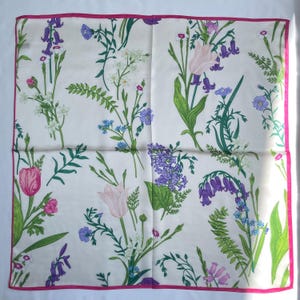 May include: A square silk scarf with a cream background featuring a botanical print of various wildflowers including purple bluebells, pink tulips, and green ferns, finished with a thin, vibrant pink border along all four edges of the fabric.
