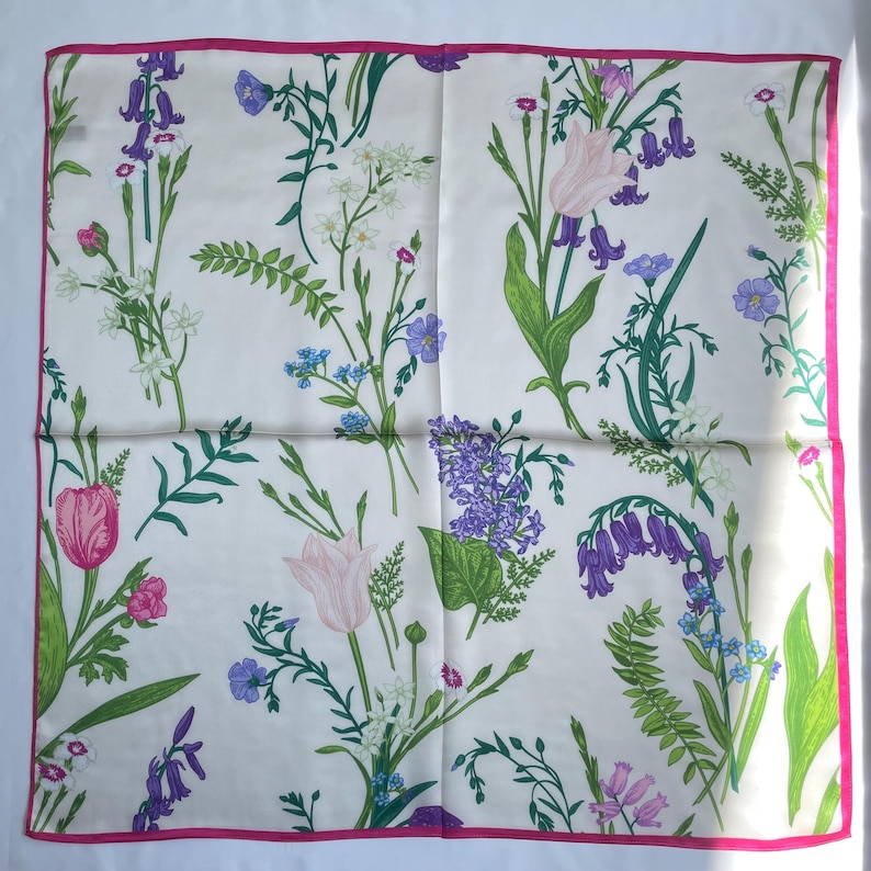 May include: A square silk scarf with a cream background featuring a botanical print of various wildflowers including purple bluebells, pink tulips, and green ferns, finished with a thin, vibrant pink border along all four edges of the fabric.