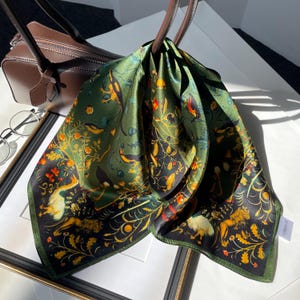 Könnte beinhalten: Ein quadratisches Seidentuch mit dunkelgrünem und schwarzem Hintergrund, bedruckt mit detaillierten Motiven von Vögeln, Löwen und goldenen Blumenmustern. Das Tuch hat eine glatte, glänzende Oberfläche und liegt über dem Griff einer braunen Ledertasche.