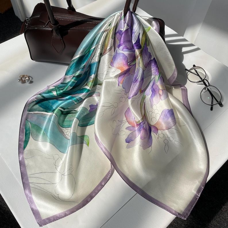 May include: A square silk scarf featuring a watercolor-style floral print with purple irises and teal green foliage on a cream background, finished with a thin lavender border. The scarf is displayed alongside a dark brown handbag and glasses.