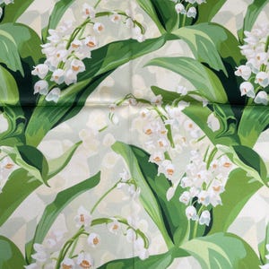 K&ouml;nnte beinhalten: Ein Stoff mit einem sich wiederholenden botanischen Muster aus wei&szlig;en Maigl&ouml;ckchen mit kleinen glockenf&ouml;rmigen Bl&uuml;ten und langen, spitzen gr&uuml;nen Bl&auml;ttern auf einem hellen, cremewei&szlig;en Hintergrund mit einer glatten, leicht gl&auml;nzenden Oberfl&auml;che.