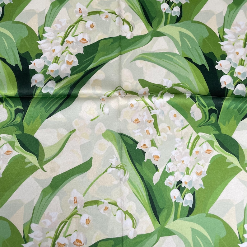 K&ouml;nnte beinhalten: Ein Stoff mit einem sich wiederholenden botanischen Muster aus wei&szlig;en Maigl&ouml;ckchen mit kleinen glockenf&ouml;rmigen Bl&uuml;ten und langen, spitzen gr&uuml;nen Bl&auml;ttern auf einem hellen, cremewei&szlig;en Hintergrund mit einer glatten, leicht gl&auml;nzenden Oberfl&auml;che.