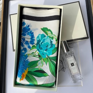 K&ouml;nnte beinhalten: Eine rechteckige wei&szlig;e Geschenkbox mit einem gefalteten Seidenschal, der ein lebhaftes Blumenmuster in Blau, T&uuml;rkis und Gr&uuml;n mit einem schwarzen Rand aufweist. Neben der Box steht ein durchsichtiger Glasflakon mit Jo Malone K&ouml;lnisch Wasser.