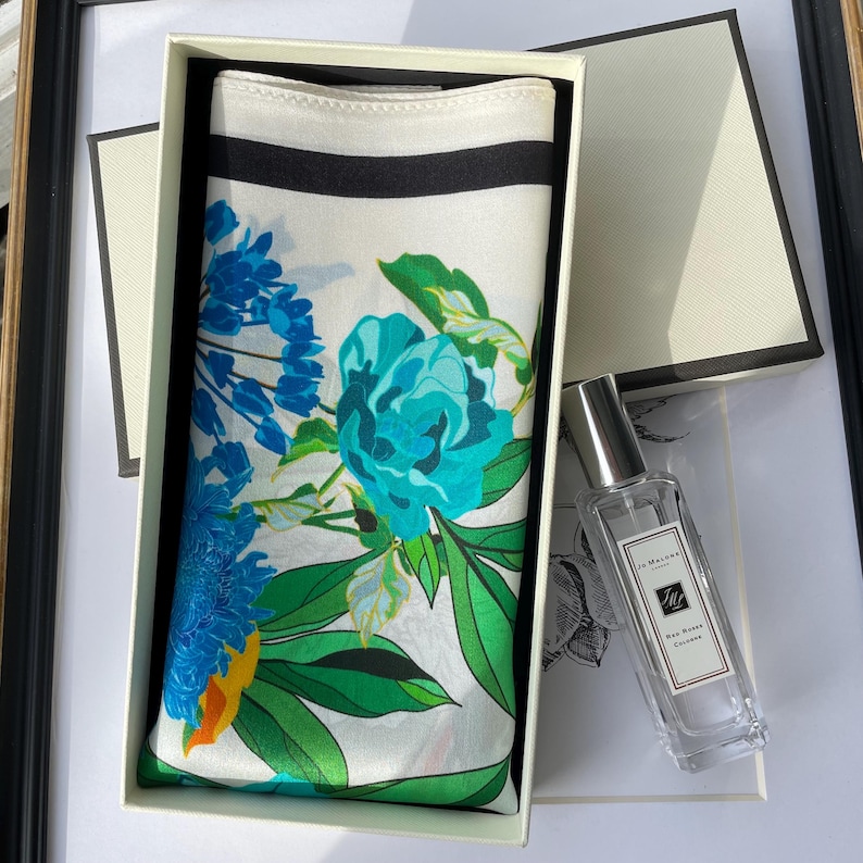 K&ouml;nnte beinhalten: Eine rechteckige wei&szlig;e Geschenkbox mit einem gefalteten Seidenschal, der ein lebhaftes Blumenmuster in Blau, T&uuml;rkis und Gr&uuml;n mit einem schwarzen Rand aufweist. Neben der Box steht ein durchsichtiger Glasflakon mit Jo Malone K&ouml;lnisch Wasser.
