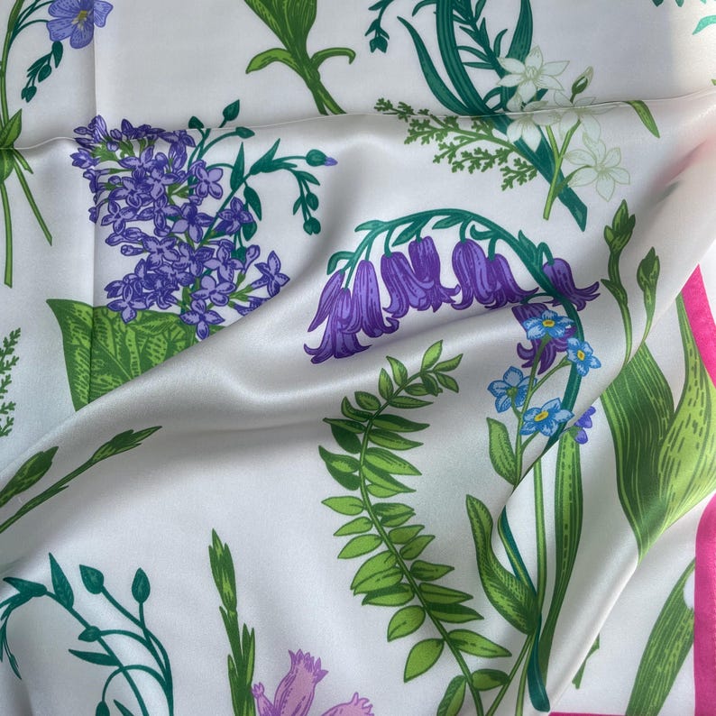 May include: A white silk scarf featuring a botanical print of purple bell-shaped flowers, clusters of small blue blossoms, and green foliage. The fabric has a smooth, reflective sheen with a bright pink border visible along the right edge.