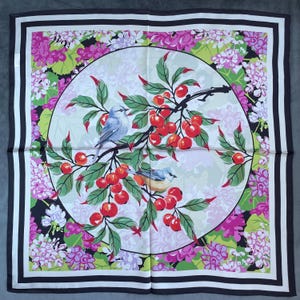 May include: A square silk scarf featuring a central circular medallion with two small birds perched on a branch laden with bright red cherries and green leaves. The background is filled with pink and purple floral clusters and a black border.