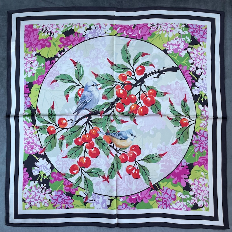 May include: A square silk scarf featuring a central circular medallion with two small birds perched on a branch laden with bright red cherries and green leaves. The background is filled with pink and purple floral clusters and a black border.