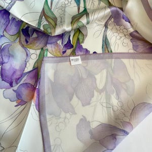 May include: A white silk scarf featuring a watercolor-style floral print with large purple irises, green stems, and delicate line-drawn foliage. A small white rectangular tag is attached to the fabric, displaying the text 90x90cm and 100% silk.