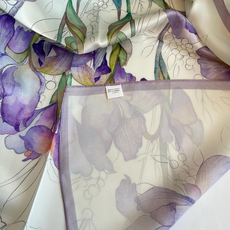 May include: A white silk scarf featuring a watercolor-style floral print with large purple irises, green stems, and delicate line-drawn foliage. A small white rectangular tag is attached to the fabric, displaying the text 90x90cm and 100% silk.