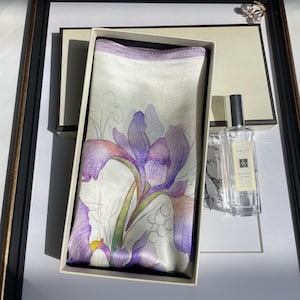 May include: A rectangular silk scarf featuring a purple and white iris flower print with a light purple border, presented inside a cream-colored gift box next to a clear glass bottle of Jo Malone Red Roses cologne with a silver cap.