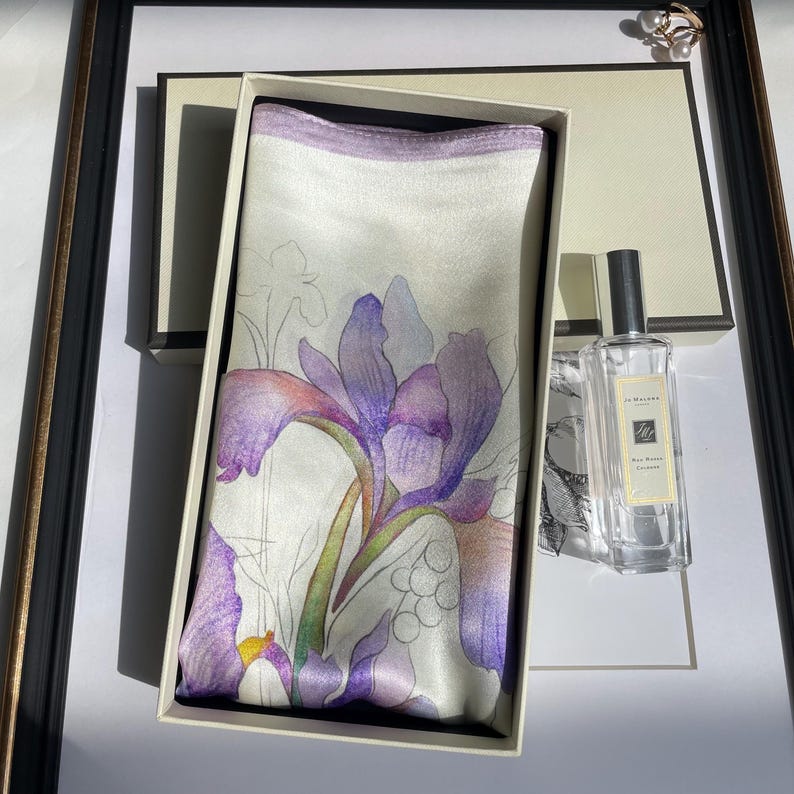 May include: A rectangular silk scarf featuring a purple and white iris flower print with a light purple border, presented inside a cream-colored gift box next to a clear glass bottle of Jo Malone Red Roses cologne with a silver cap.