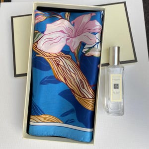 May include: A square silk scarf featuring a vibrant blue background with large pink floral illustrations and golden brown leaf patterns, presented in a rectangular cream gift box alongside a clear glass bottle of Jo Malone Red Roses cologne.