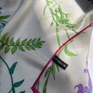 May include: A white silk scarf featuring a botanical print of green fern leaves, white flowers, and purple floral accents. The scarf has a bright pink stitched edge and a black fabric tag that reads 100% Mulberry Silk in white text.