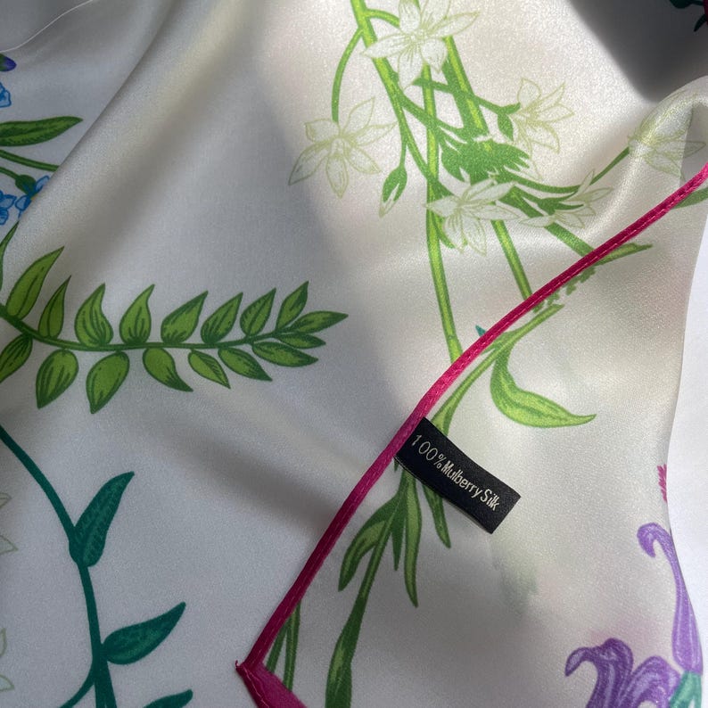 May include: A white silk scarf featuring a botanical print of green fern leaves, white flowers, and purple floral accents. The scarf has a bright pink stitched edge and a black fabric tag that reads 100% Mulberry Silk in white text.
