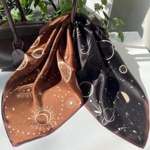 Könnte beinhalten: Ein quadratisches Seidentuch mit einem geteilten Design in Braun und Schwarz. Der Stoff ist mit weißen Himmelsmotiven bedruckt, darunter Sonnen, Monde, Planeten, Sterne und Sternbilder, die über die gesamte Oberfläche verteilt sind.