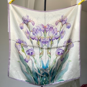 May include: A square silk scarf featuring a white background with a central botanical illustration of purple and blue iris flowers. The design includes green stems and leaves, with a light purple border framing the entire square fabric piece.