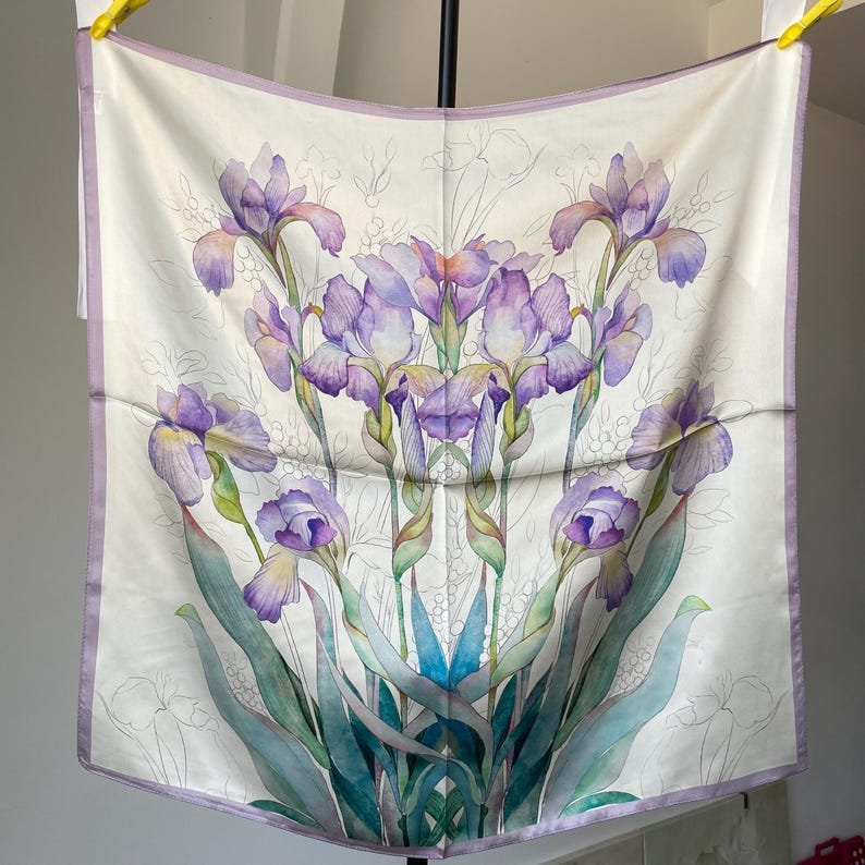 May include: A square silk scarf featuring a white background with a central botanical illustration of purple and blue iris flowers. The design includes green stems and leaves, with a light purple border framing the entire square fabric piece.